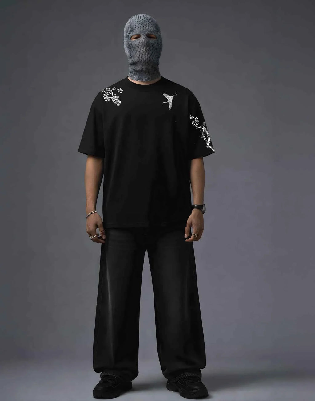 Black graphic tee with white crane and floral embroidery, paired with wide-leg pants and knit balaclava for edgy streetwear style.